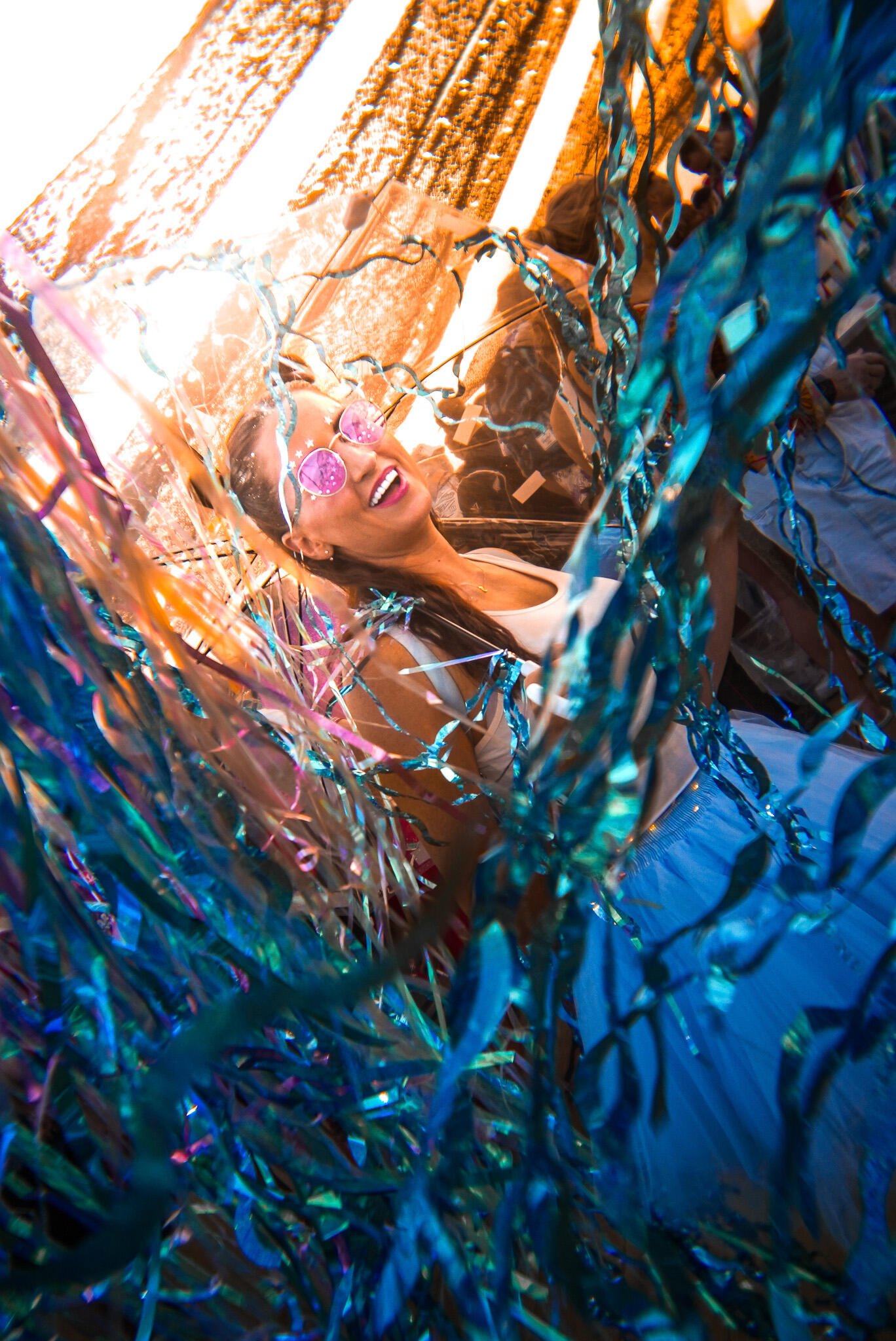 Rugby7s Dubai Woman in pink sunglasses smiling and dancing through metallic streamers at an outdoor festival, lit by warm sunlight.