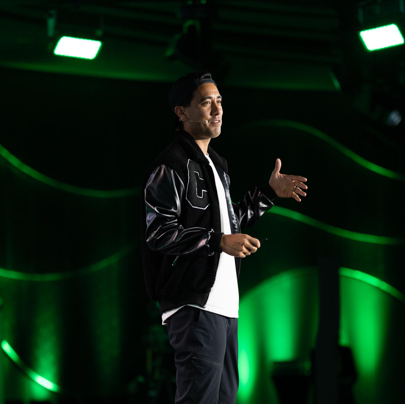 CCCC Dubai Man wearing a black varsity jacket, white t-shirt, and red sneakers speaks on stage under green lighting at a professional event. He gestures with one hand and wears a headset microphone.
